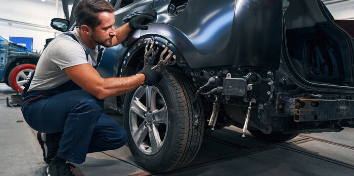 Concentrated light vehicle technician attaching screw clamps to car frame above rear wheel while flattening its surface during auto body repair